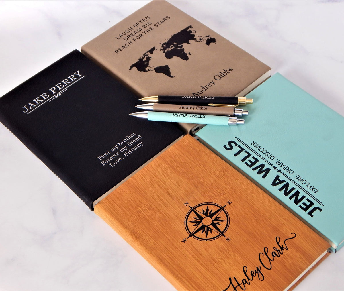 Four personalized hardcover notebooks in black, tan, light blue, and natural bamboo wood, each with a matching pen, arranged on a white marble surface. The notebooks feature custom names, travel-themed quotes, and black printed designs including a world map and compass rose.