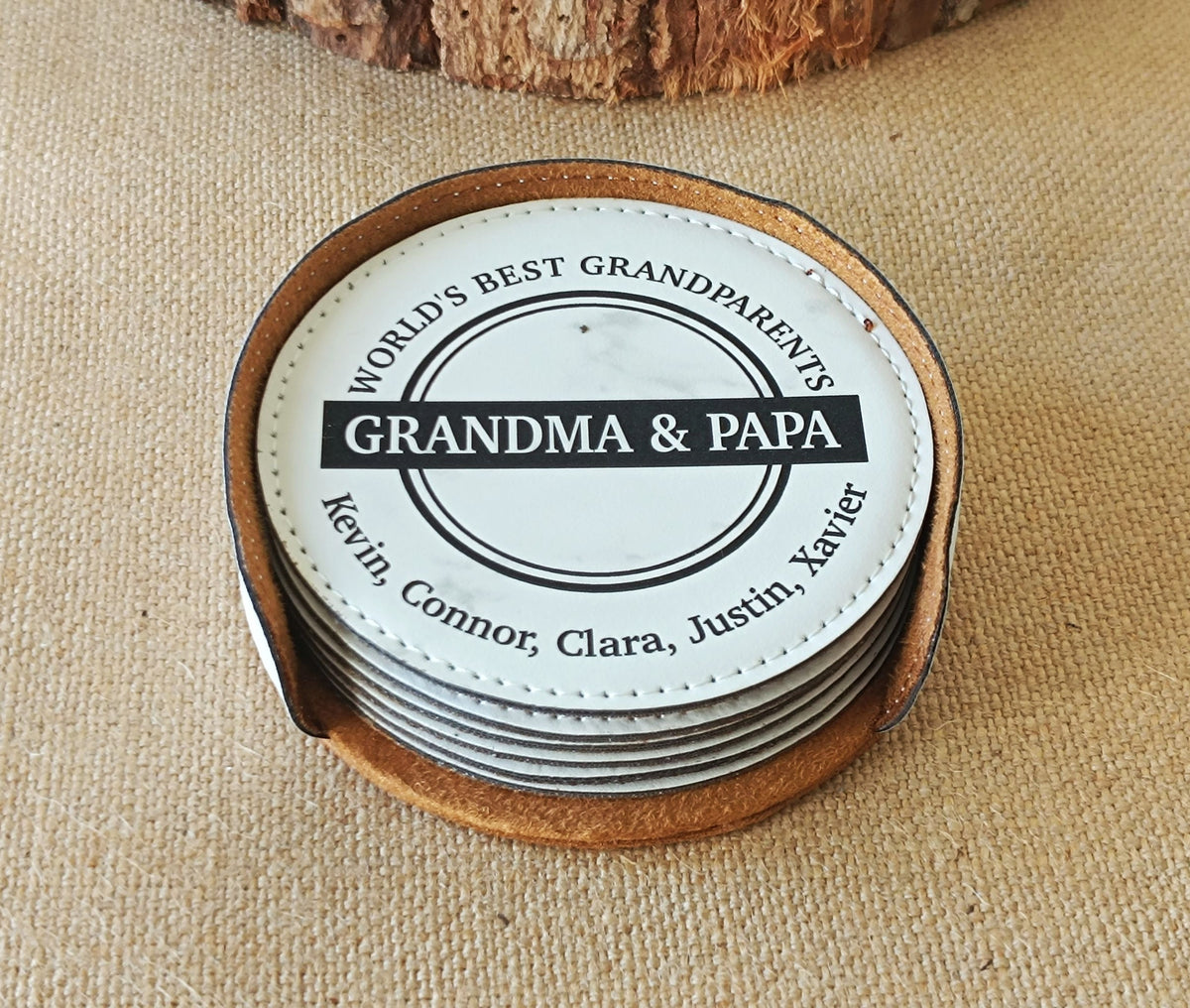 A set of round dark gray coasters stacked together, featuring a top coaster with a personalized design that reads 'WE LOVE You GRANDMA' with a heart and the names 'Sherry' and 'Juan', surrounded by a tan leather trim on a white background.