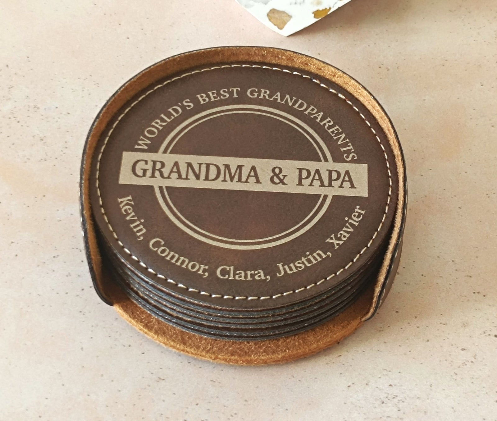 A stack of personalized leather coasters on a light beige speckled countertop. The top coaster features a circular design with a dark brown center, cream-colored text reading 'WORLD'S BEST GRANDPARENTS GRANDMA & PAPA Kevin, Connor, Clara, Justin, Xavier', and a stitched leather border.