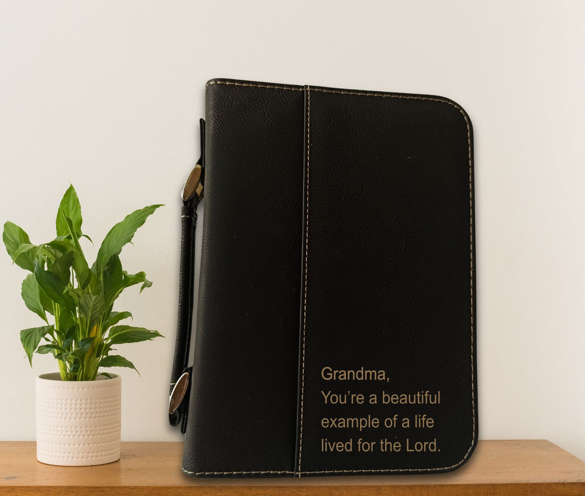 A black faux-leather Bible cover or tablet sleeve with gold text reading 'Grandma, You're a beautiful example of a life lived for the Lord.' sits on a wooden shelf next to a potted green plant against a plain off-white wall.