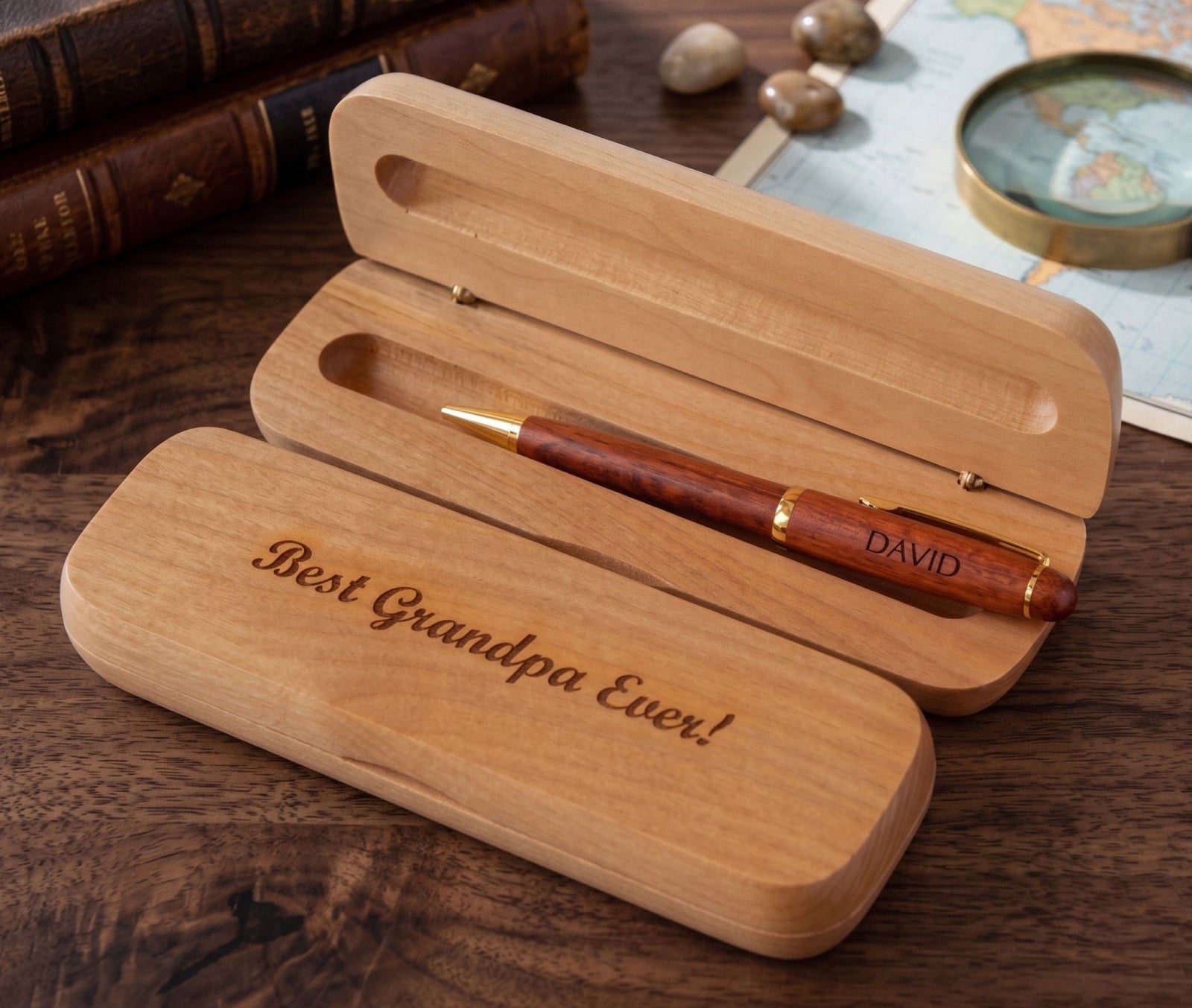A personalized wooden pen gift set on a dark wood tabletop, featuring a wooden case engraved with 'Best Grandpa Ever!' and a matching wooden pen with gold trim and the name 'DAVID' on the barrel.