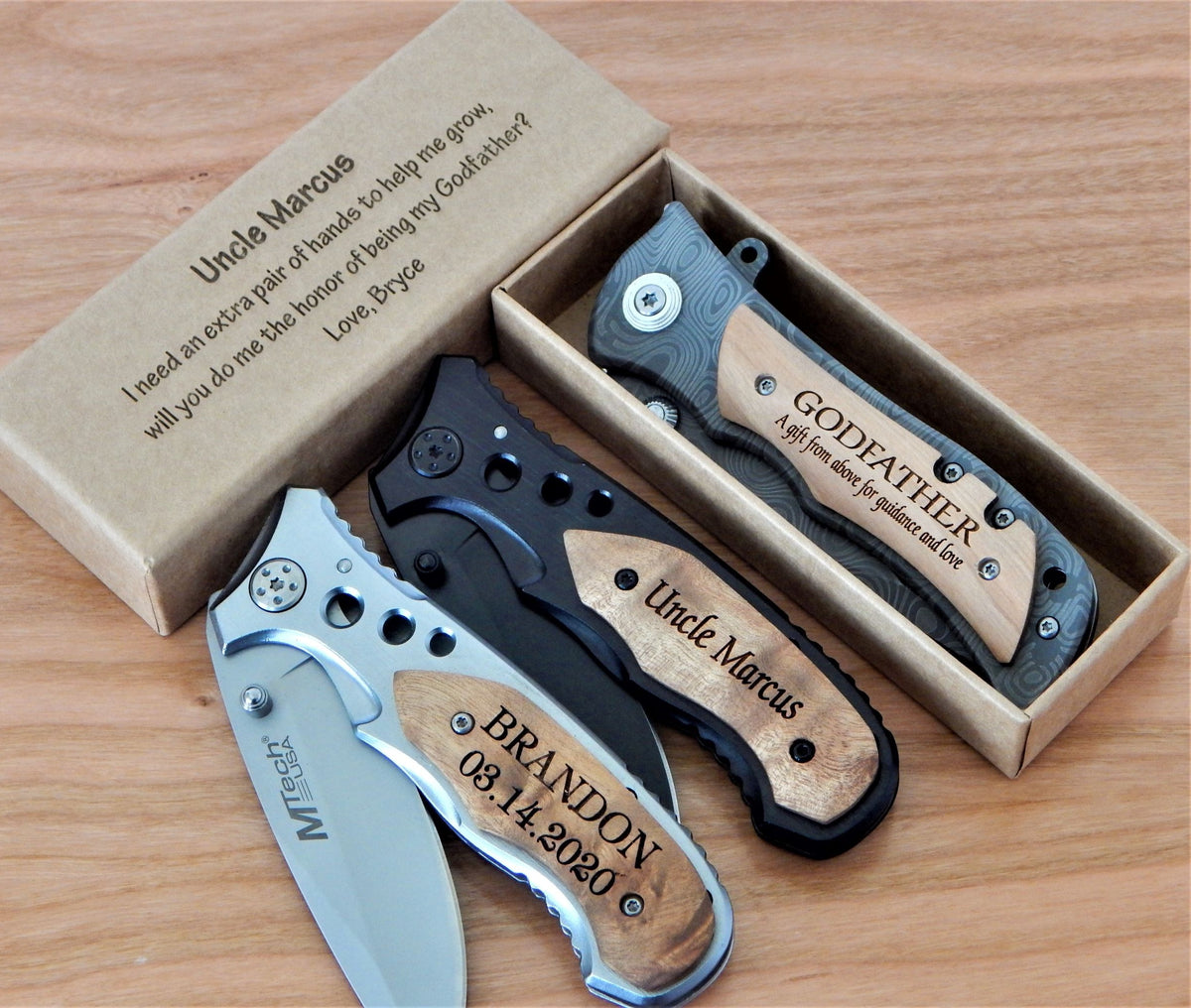 Three personalized folding pocket knives displayed on a wooden surface with their open and closed views, alongside a kraft paper gift box printed with a message for 'Uncle Marcus'. The knives feature wood-grain handles with laser-engraved names and dates, and one has a silver blade.