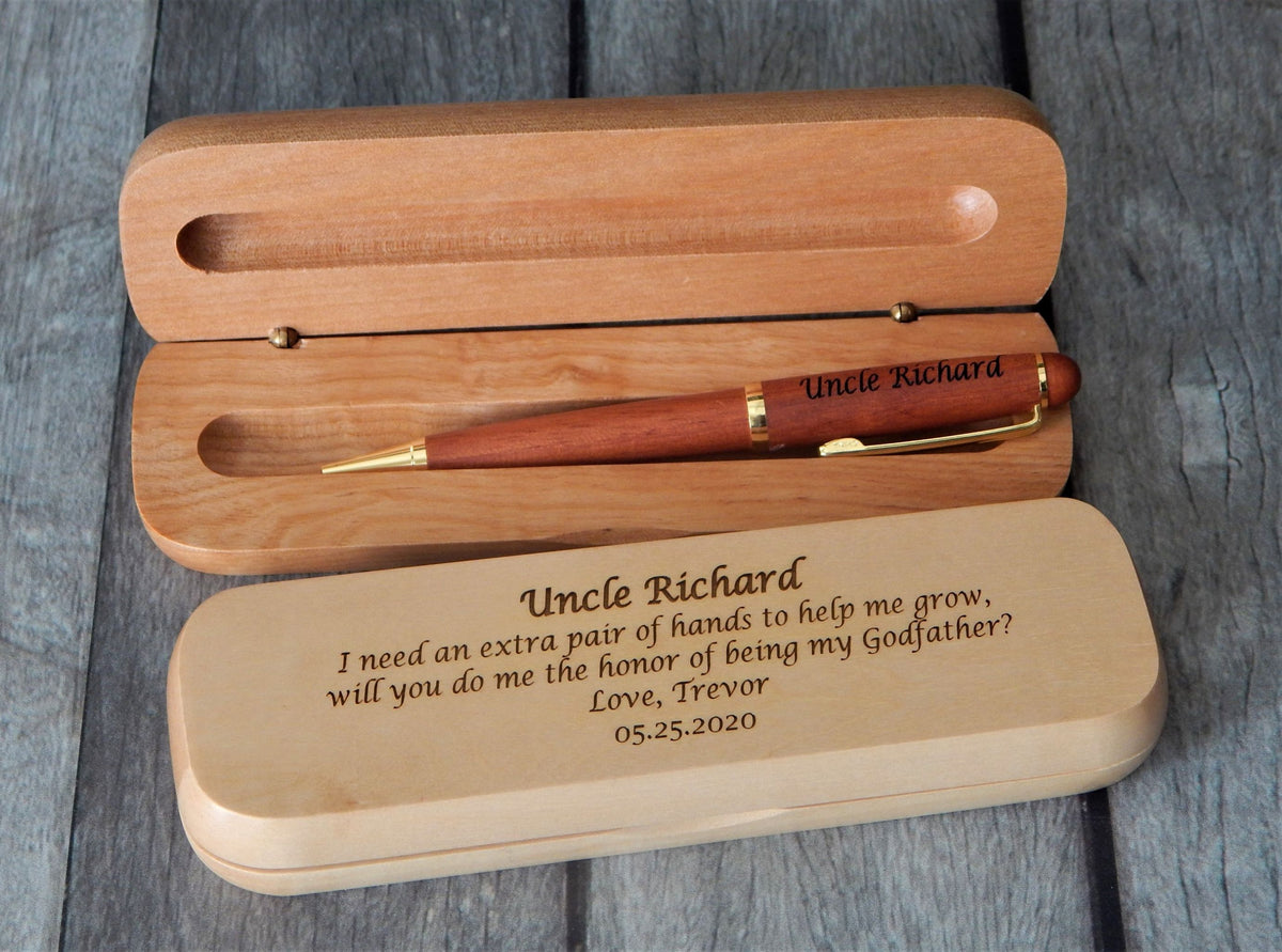 Personalized wooden keepsake box set with an engraved pen, shown on a gray wood plank background. The set includes an open light brown wooden box with a reddish-brown pen inside, and a closed matching box in front engraved with a message for 'Uncle Richard'.