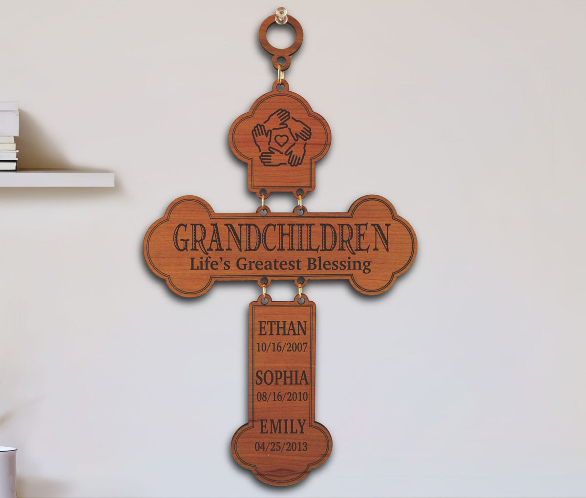 A wooden hanging cross-shaped memorial plaque engraved with the words “GRANDCHILDREN” and “Life’s Greatest Blessing,” with three hanging tabs below listing names and dates for Ethan, Sophia, and Emily, shown against a light wall.