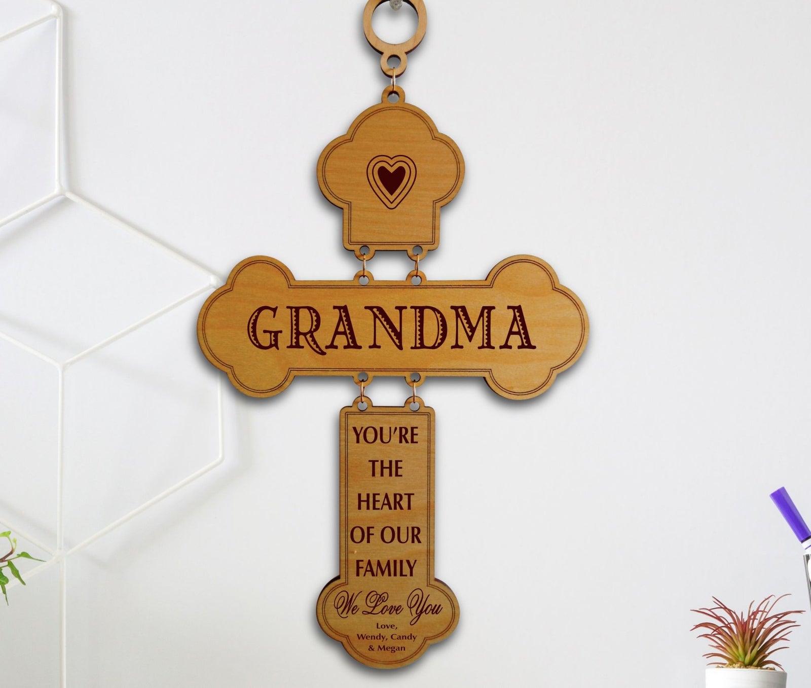 A personalized wooden cross hanging ornament engraved with 'GRANDMA' and a sentimental message, displayed against a white wall with subtle home decor elements.