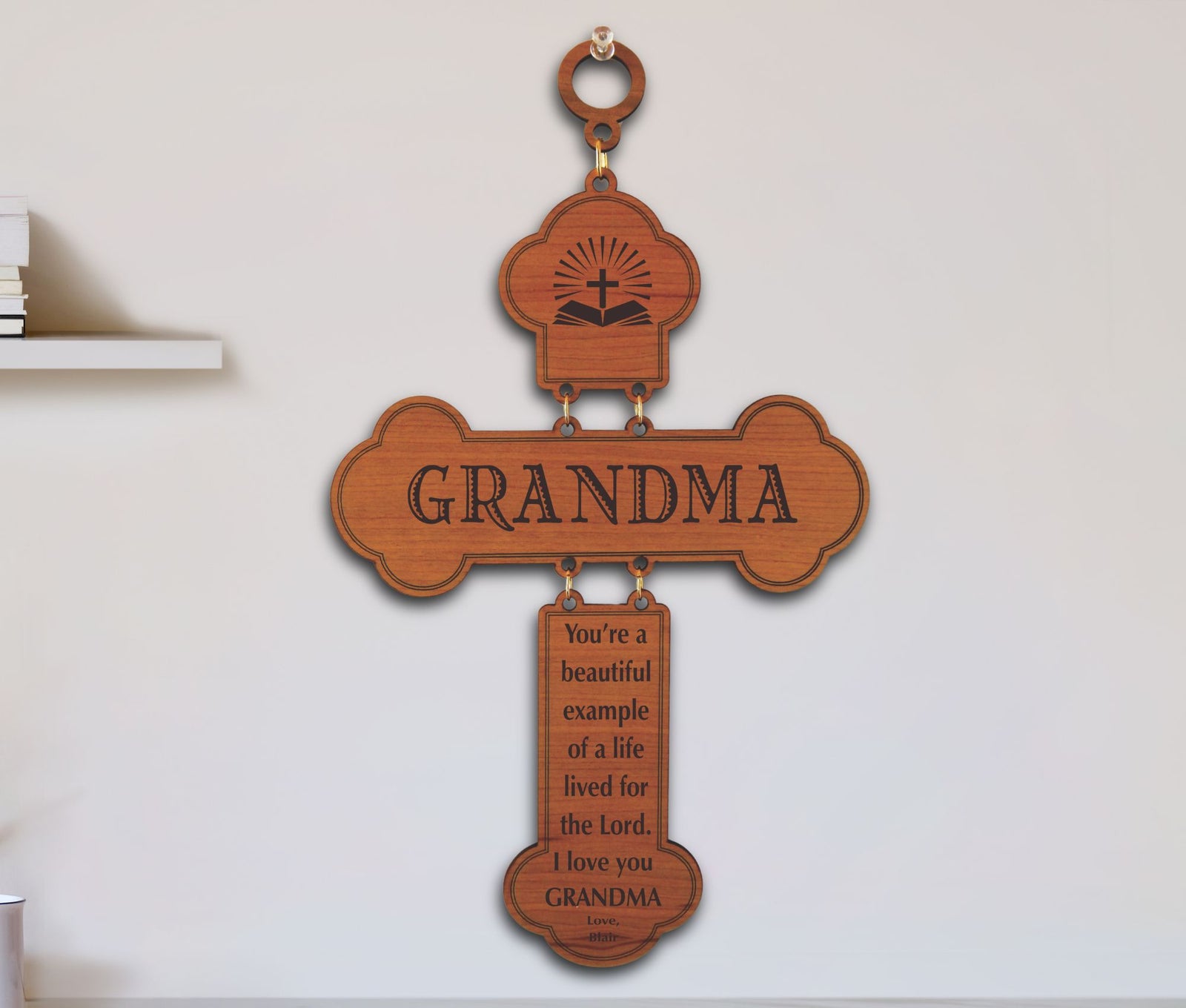 A wooden cross-shaped wall hanging with the word GRANDMA across the center and an engraved Bible verse below, mounted on a light wall.
