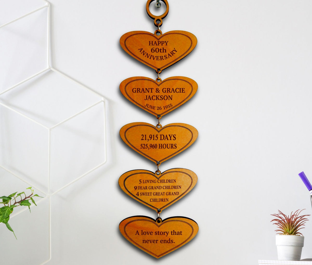 A vertical wooden anniversary wall hanging with five engraved heart-shaped plaques connected by small metal rings, shown hanging on a white wall in a styled interior setting.
