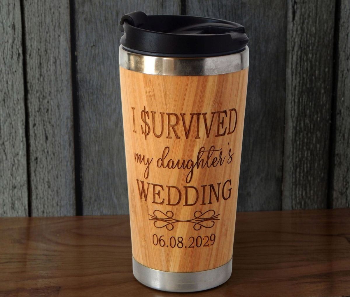 A bamboo wood-look insulated travel tumbler with a black lid, featuring the engraved text 'I SURVIVED my daughter's WEDDING 06.08.