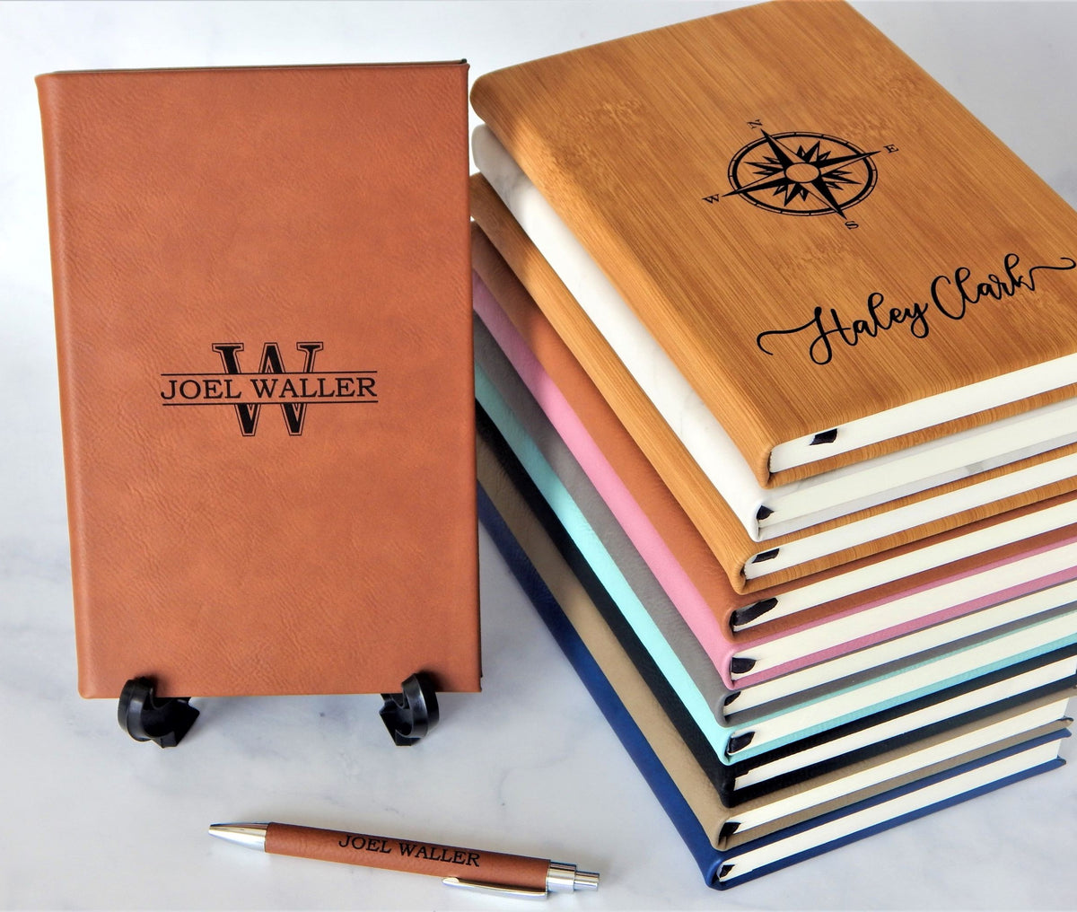 A stack of personalized hardcover notebooks and a matching pen on a light marble surface. The stack includes bamboo, cream, pink, blue, and navy covers, with the top notebook engraved with a compass and the name 'Harley Clark'.