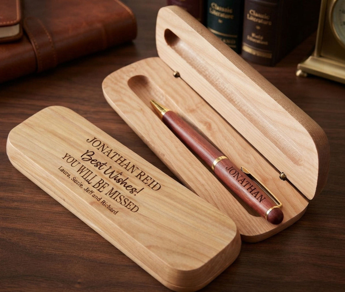 A personalized wooden pen case shown open on a dark wood desk, with a reddish-brown pen resting inside. The engraved lid reads 'JONATHAN REID Best Wishes! YOU WILL BE MISSED' with a memorial inscription below.