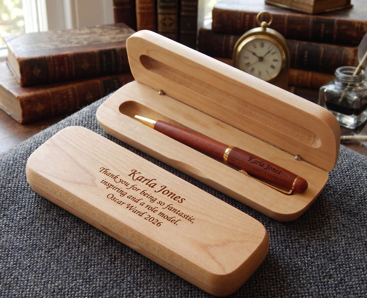 A wooden pen presentation case shown open and closed on a textured gray surface, with vintage books, a pocket watch, and a small glass bottle in the background. The cases are engraved with the name Karla Jones, and a reddish-brown pen with gold trim rests inside the open case.