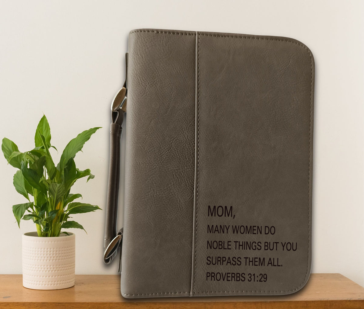 A gray faux-leather Bible cover with a zipper closure and black printed text reading 'MOM, MANY WOMEN DO NOBLE THINGS BUT YOU SURPASS THEM ALL. PROVERBS 31:29', displayed on a wooden shelf next to a potted green plant against a white wall.