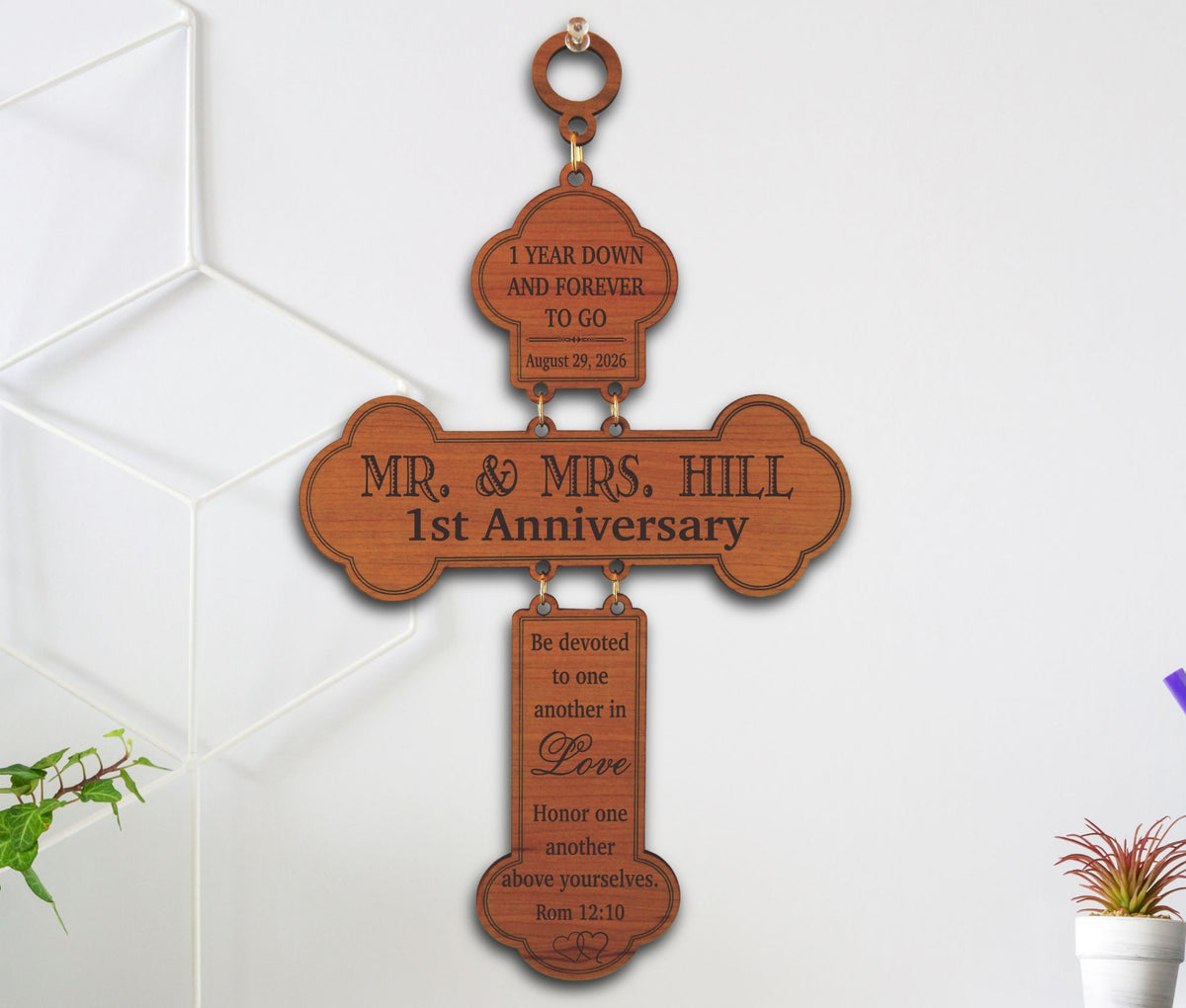A personalized wooden cross wall hanging with engraved anniversary text, shown hanging on a white wall with a geometric wall decal and small potted plants nearby.