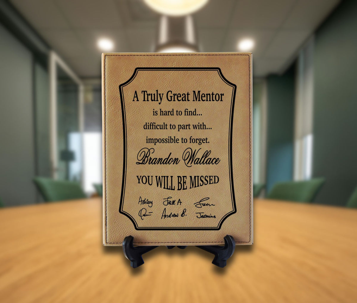 A personalized tan leather memorial plaque on a small black easel stand, displayed on a wooden table with a blurred office background.