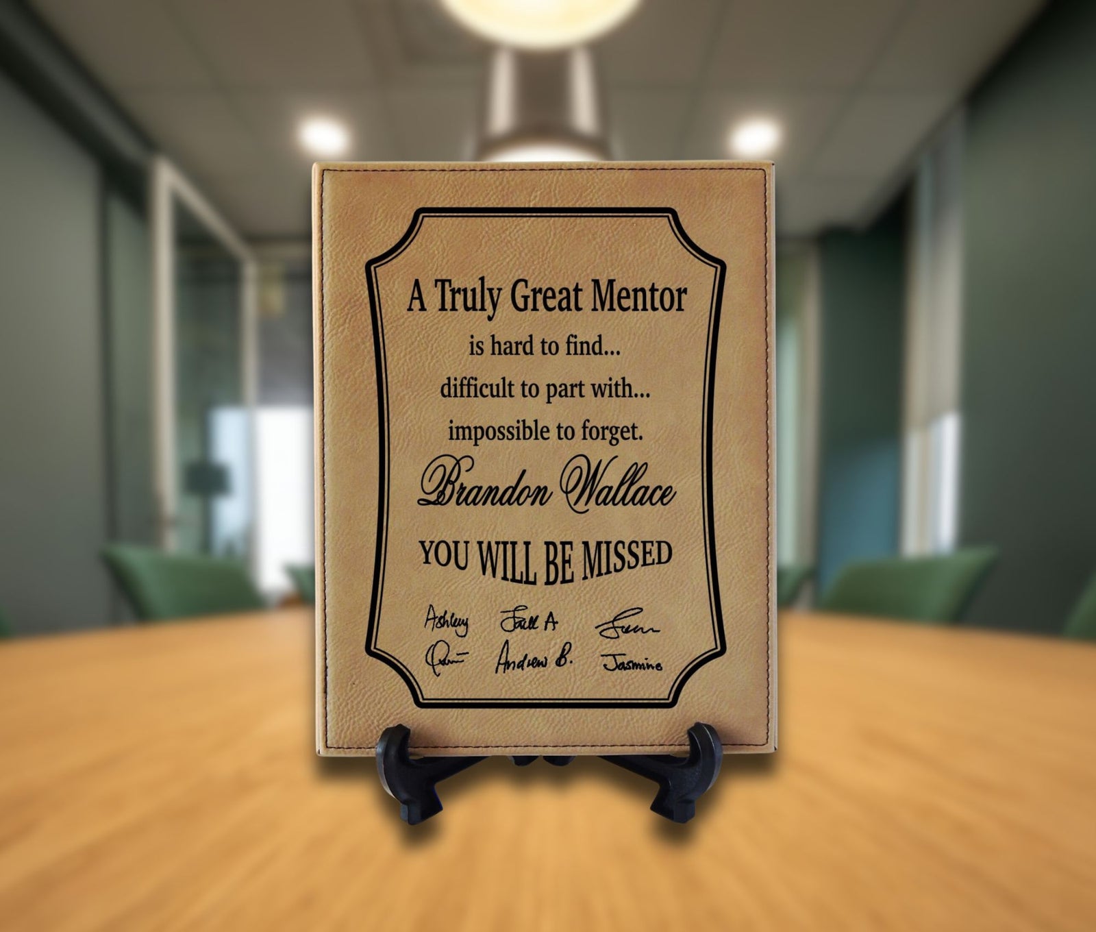 A personalized tan leather memorial plaque on a small black easel stand, displayed on a wooden table with a blurred office background.