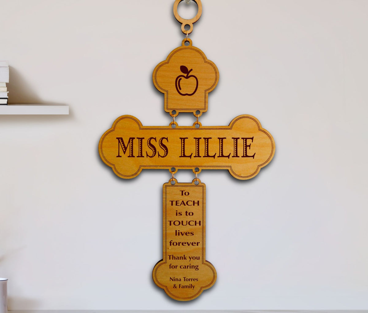 A personalized wooden cross wall plaque hanging on a white wall. The cross features an apple icon at the top, the engraved text 'MISS LILLIE' across the center, and a hanging plaque below with a thank you message for a teacher, 'To TEACH is to TOUCH lives forever.