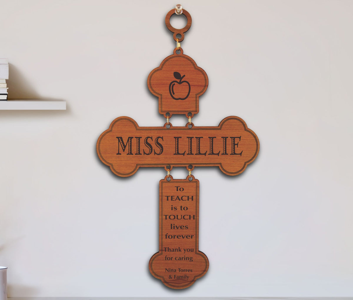 A personalized wooden cross wall hanging with an apple icon at the top, the name 'MISS LILLIE' across the center, and a hanging plaque that reads 'to TEACH is to TOUCH lives forever Thank you for caring - Nina Torres &amp; Family' on a white wall.