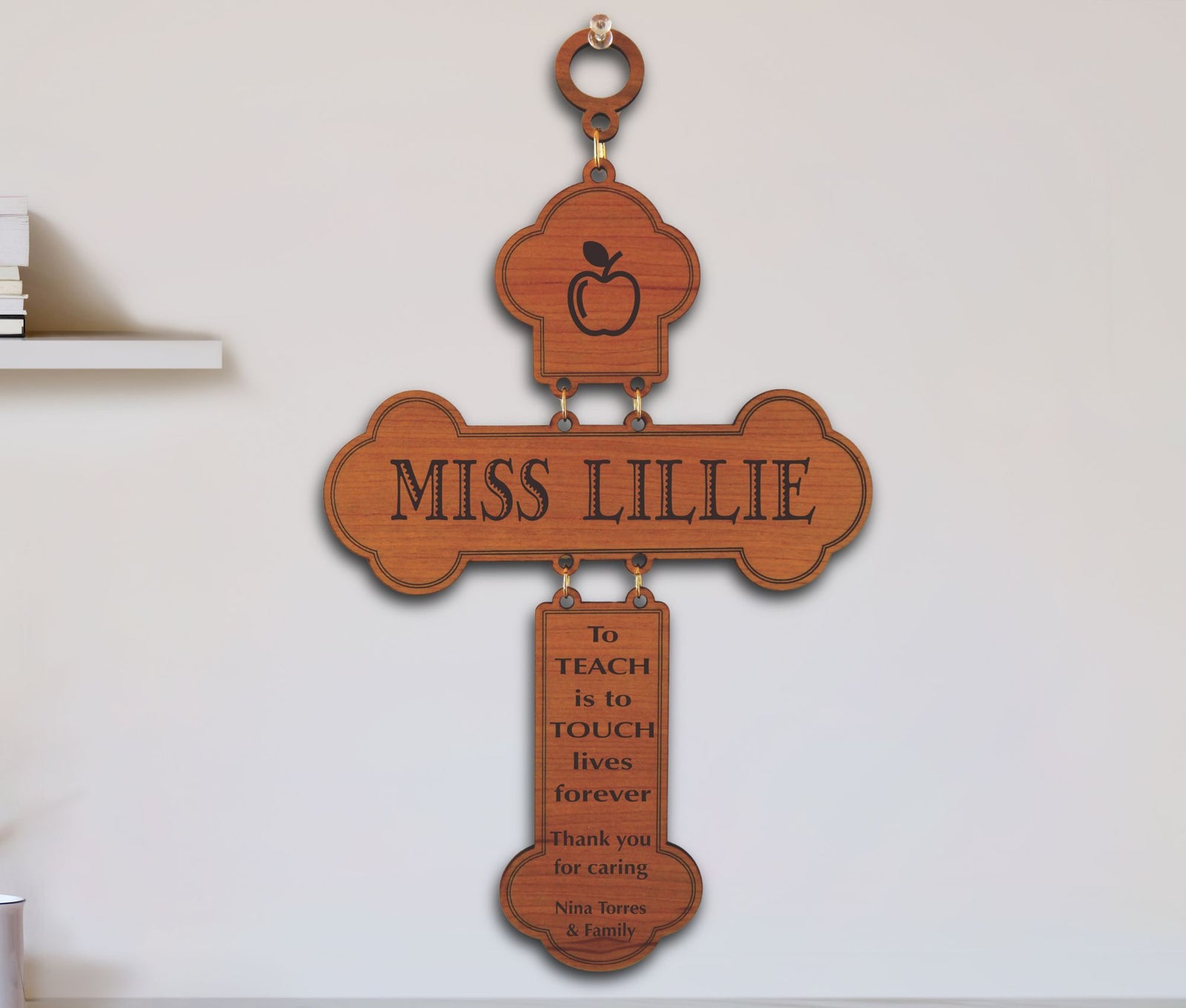 A personalized wooden cross wall hanging with an apple icon at the top, the name 'MISS LILLIE' across the center, and a hanging plaque that reads 'to TEACH is to TOUCH lives forever Thank you for caring - Nina Torres & Family' on a white wall.