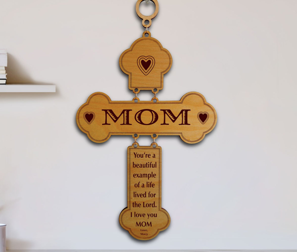 A wooden cross-shaped wall hanging with the word “MOM” engraved across the center and a smaller plaque below containing a message for Mom. The piece is shown hanging on a plain light wall.