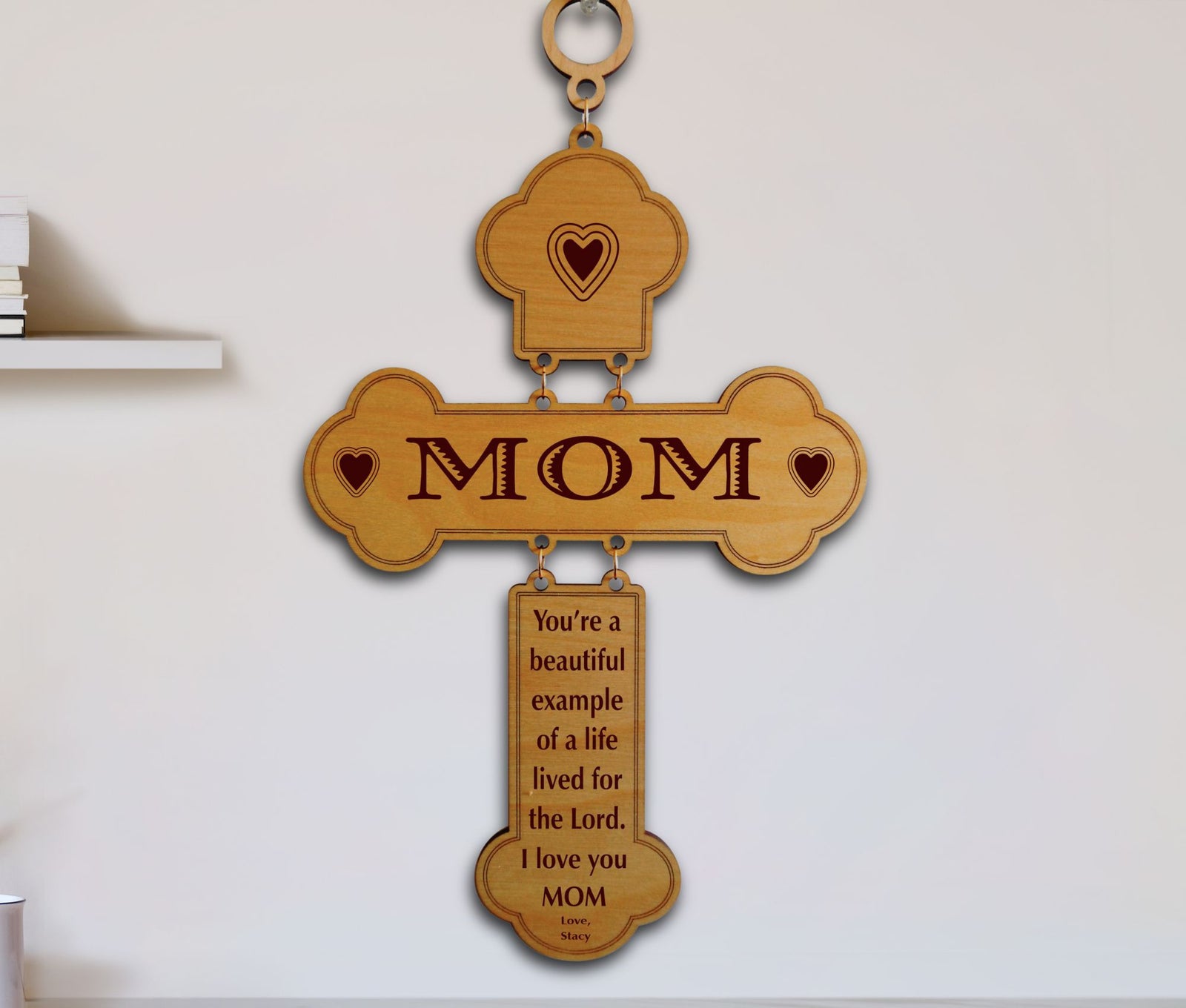 A wooden cross-shaped wall hanging with the word “MOM” engraved across the center and a smaller plaque below containing a message for Mom. The piece is shown hanging on a plain light wall.