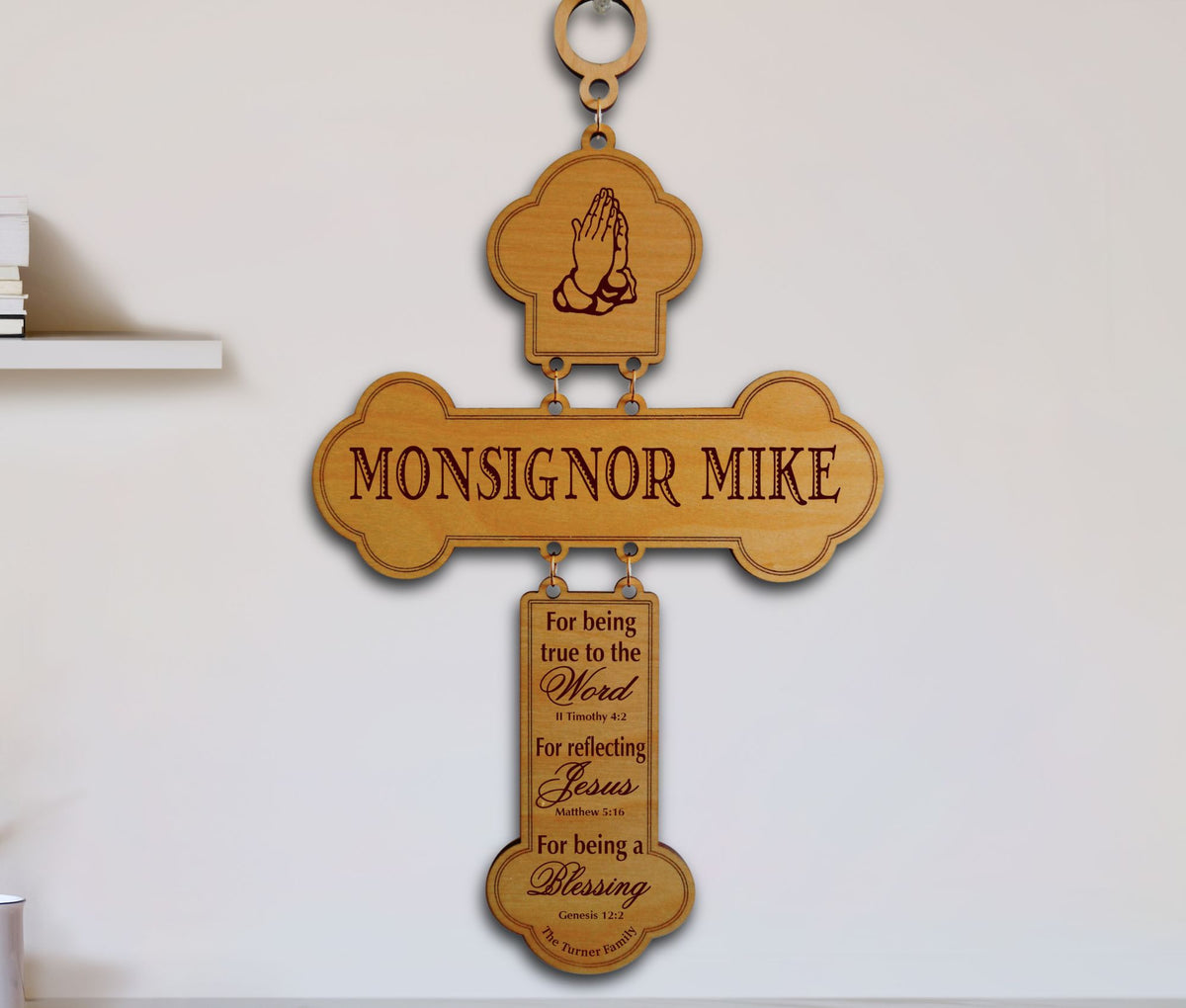 A personalized wooden cross wall hanging with a top loop, engraved with 'MONSIGNOR MIKE' and a praying hands icon, plus a hanging plaque with inspirational religious text, shown against a white wall.