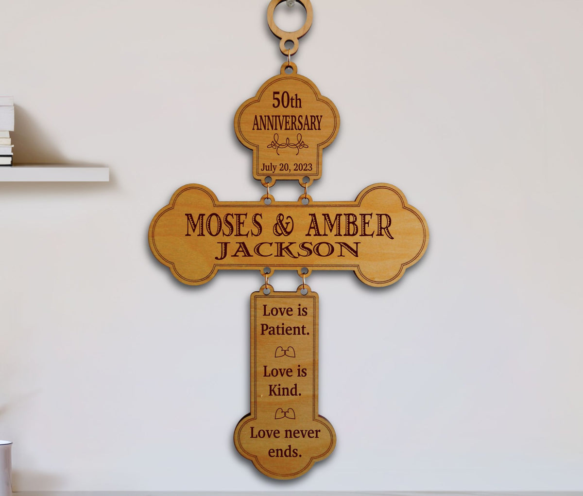 A personalized wooden cross wall plaque engraved with '50th Anniversary' and the names 'MOSES &amp; AMBER JACKSON,' plus a hanging tag reading 'Love is Patient. Love is Kind.