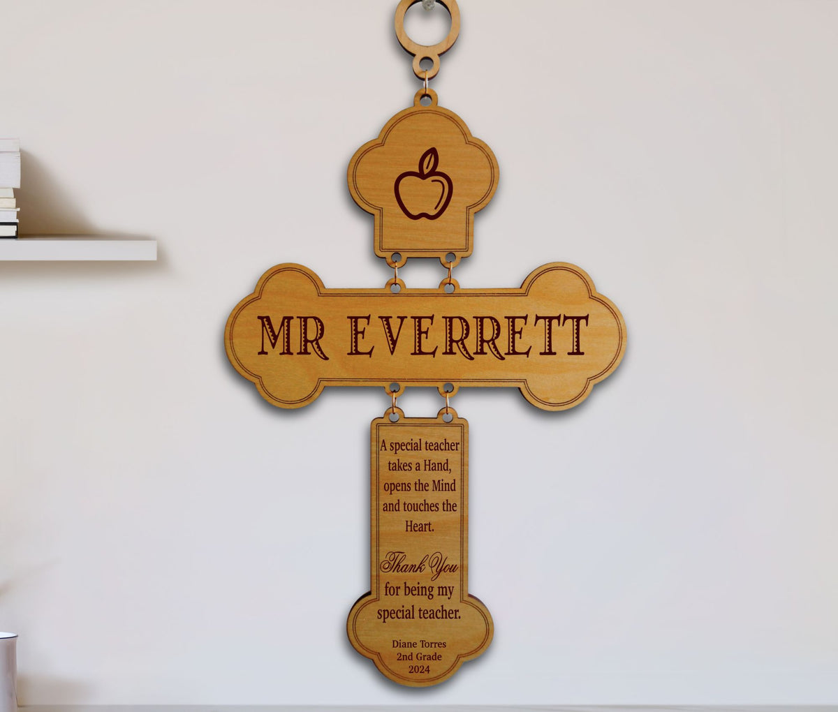 A personalized wooden cross-shaped wall plaque hanging on a white wall. The top section features an apple engraving, the center reads 'MR EVERRETT', and a hanging tag below contains a thank-you message for a special teacher, with 'Name: Everett 2nd Grade 2024' at the bottom.