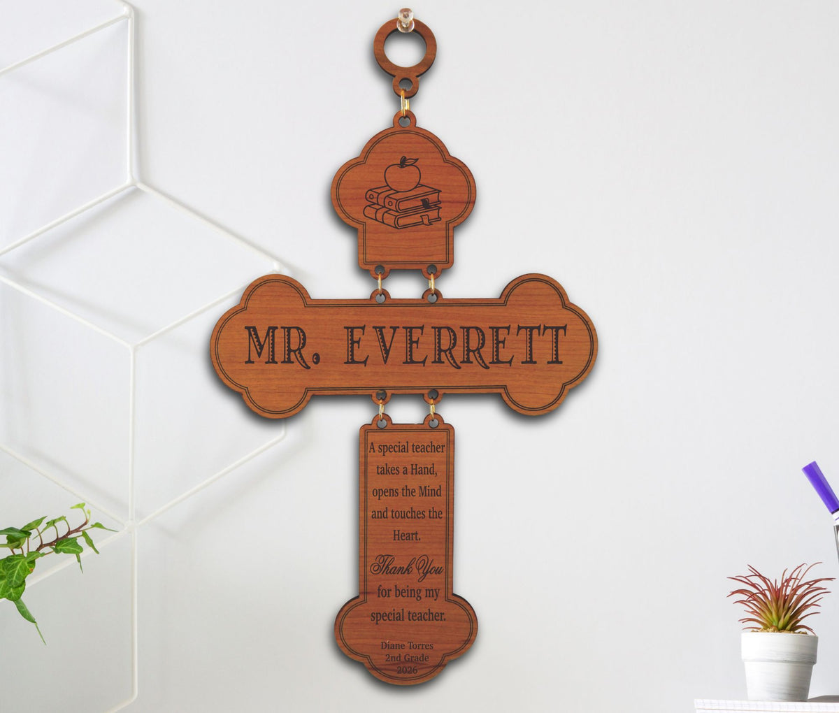 Personalized wooden cross wall plaque engraved with 'MR. EVERRETT' and a thank you message for a special teacher, hanging on a white wall with decorative plants nearby.