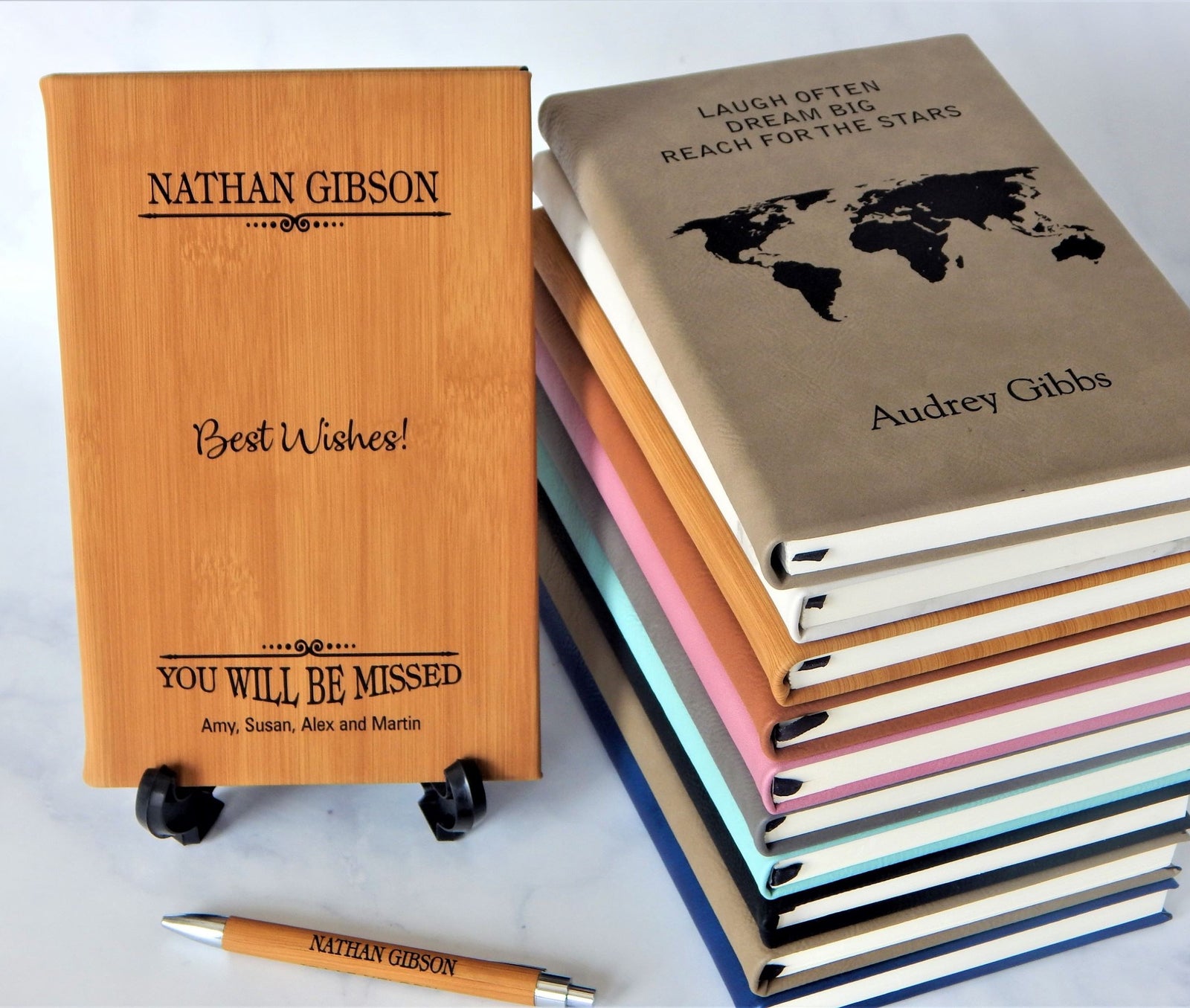 Personalized leather engraved notebooks for boss or coworker leaving displayed on a light marble surface, including one standing upright on a small stand and a stack of closed notebooks. A matching bamboo leather pen lies in front.