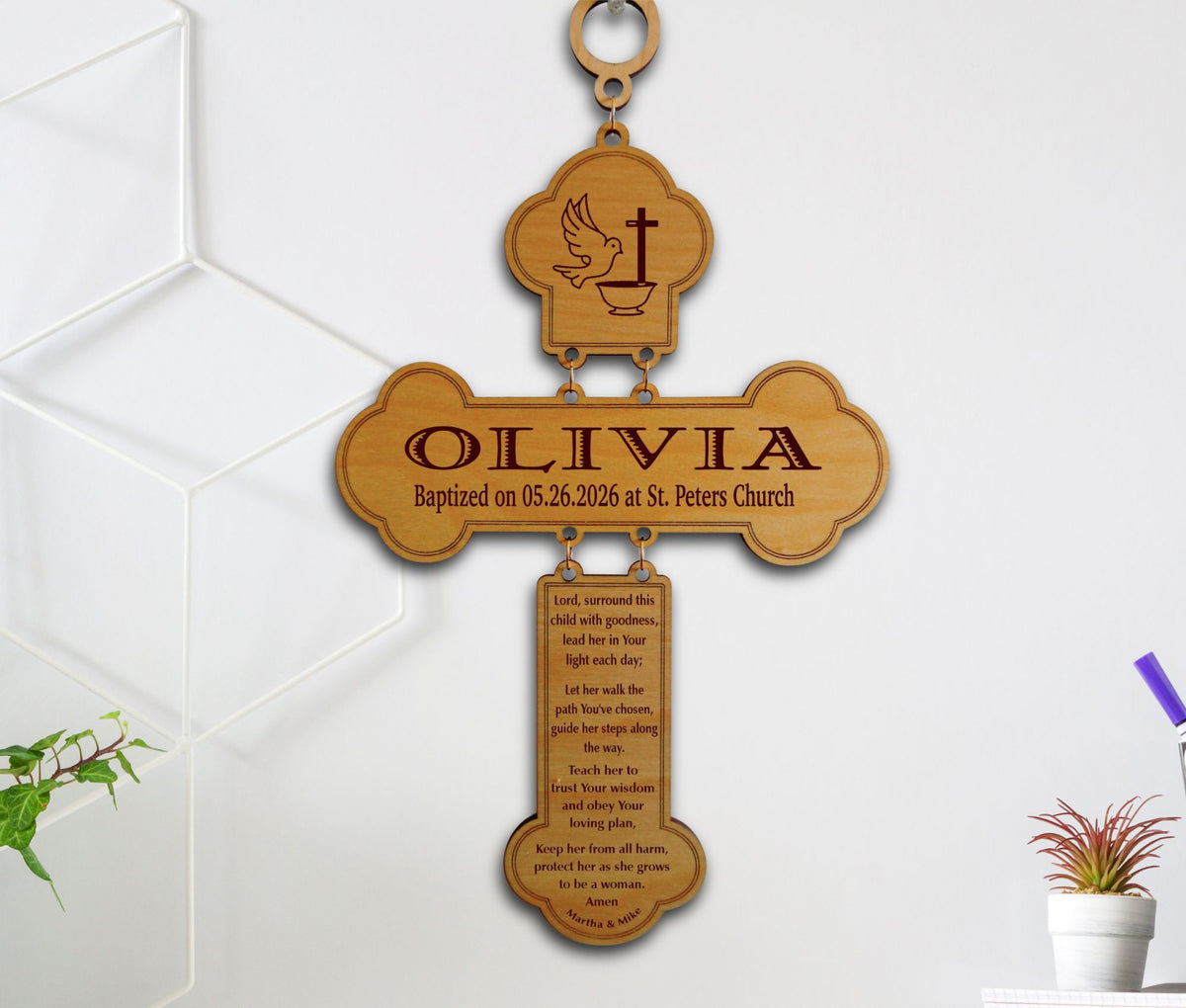 A personalized wooden baptism cross plaque hanging on a white wall. The cross is engraved with a dove and cross at the top, the name 'OLIVIA' in the center, and a longer inscription below.