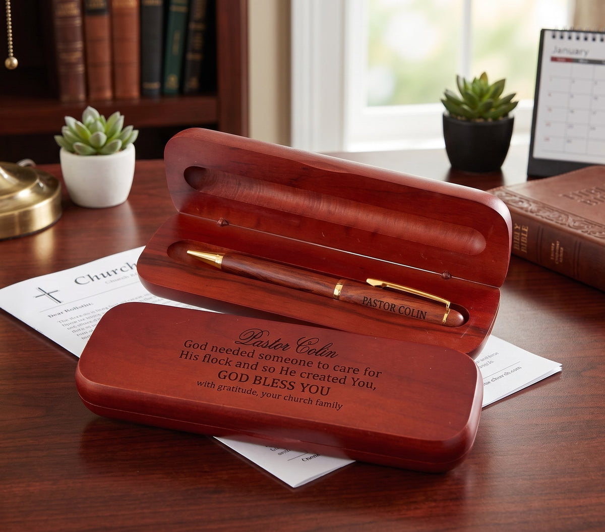 A personalized wooden keepsake box set on a wooden desk, featuring an open box with a matching wooden pen inside and a closed box in front with engraved text. The scene is styled with a book, succulents, and papers on a warm wooden table.