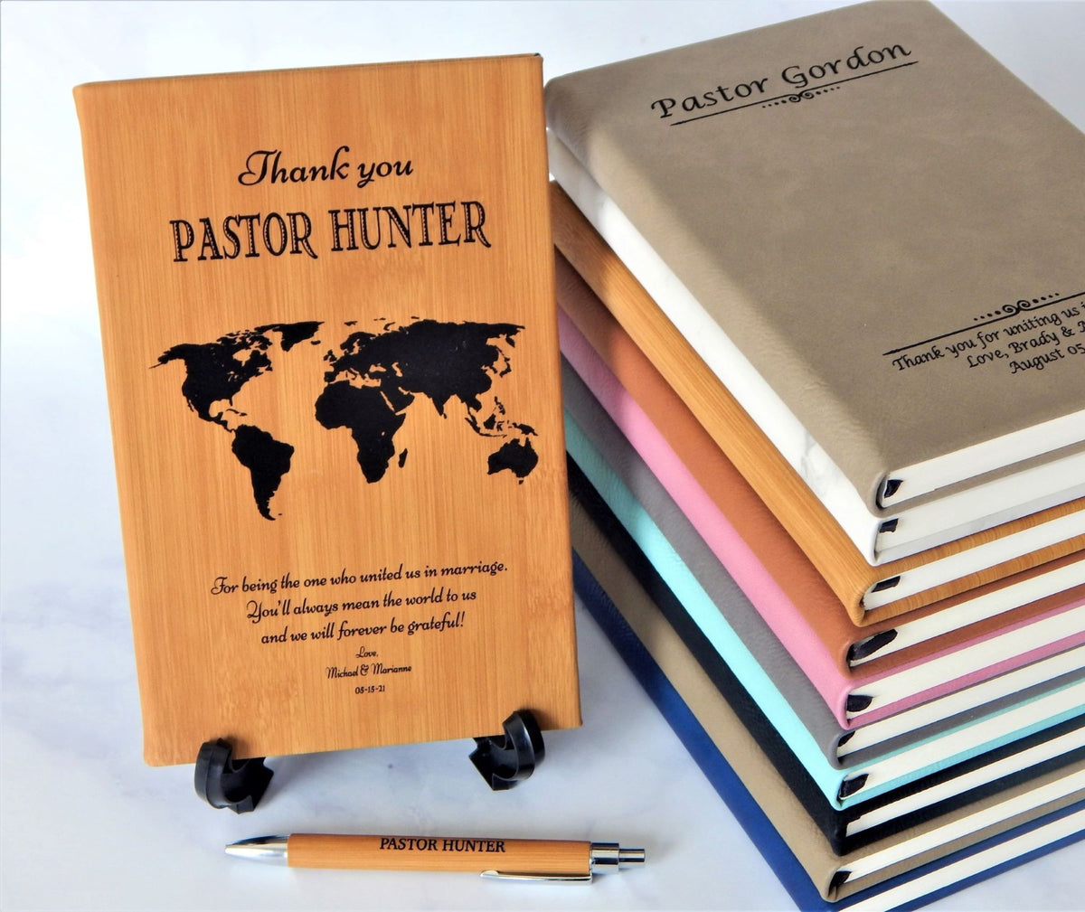 A stack of personalized wooden keepsake journals with different colored covers, with one wooden journal standing upright on a small black stand in front. The standing journal is engraved with 'Thank you PASTOR HUNTER' and a black world map graphic.