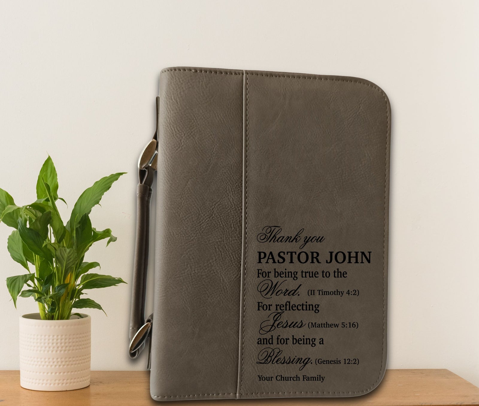 A gray faux-leather binder cover with a wrist strap, featuring black printed personalized text, displayed on a wooden surface next to a potted green plant against a white wall.