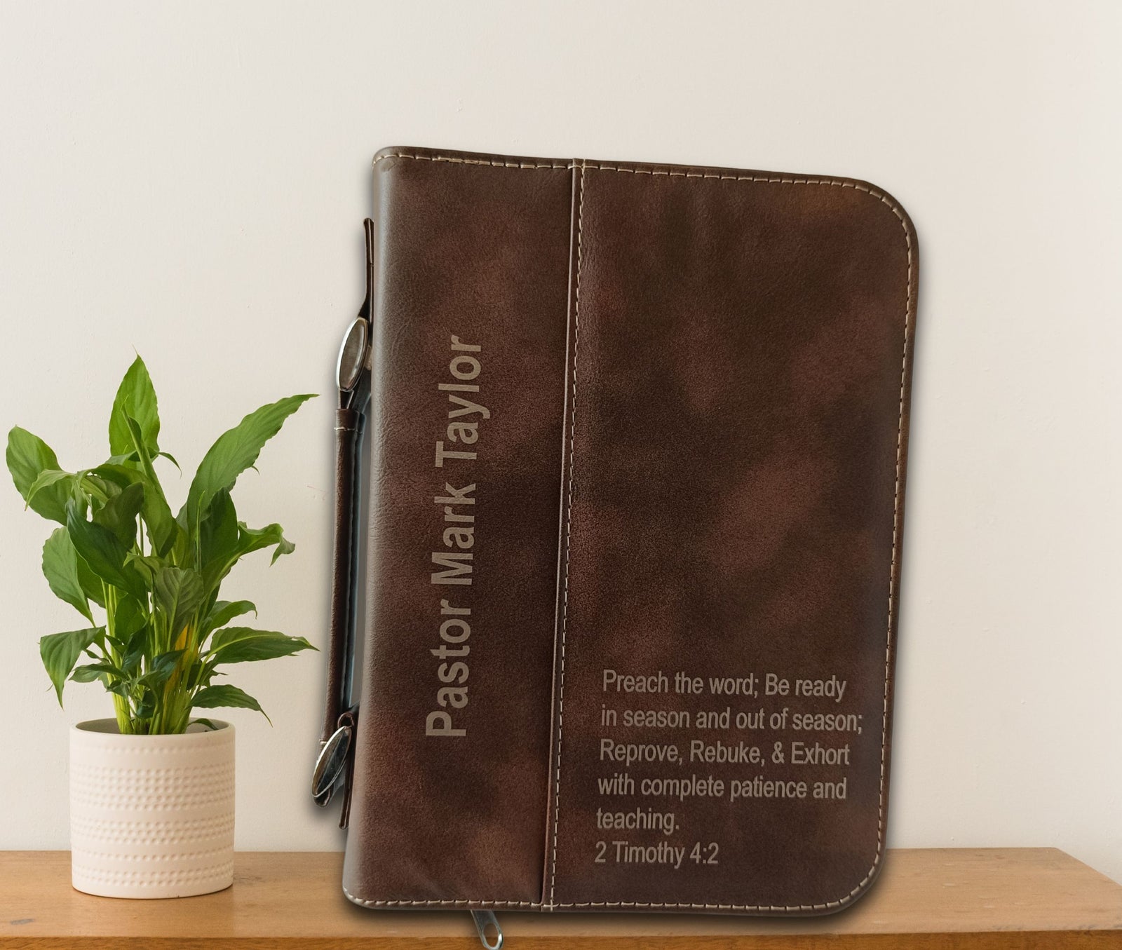 A brown leather Bible cover with custom engraving for 'Pastor Mark Taylor' sits on a wooden shelf next to a potted green plant against a plain off-white wall.