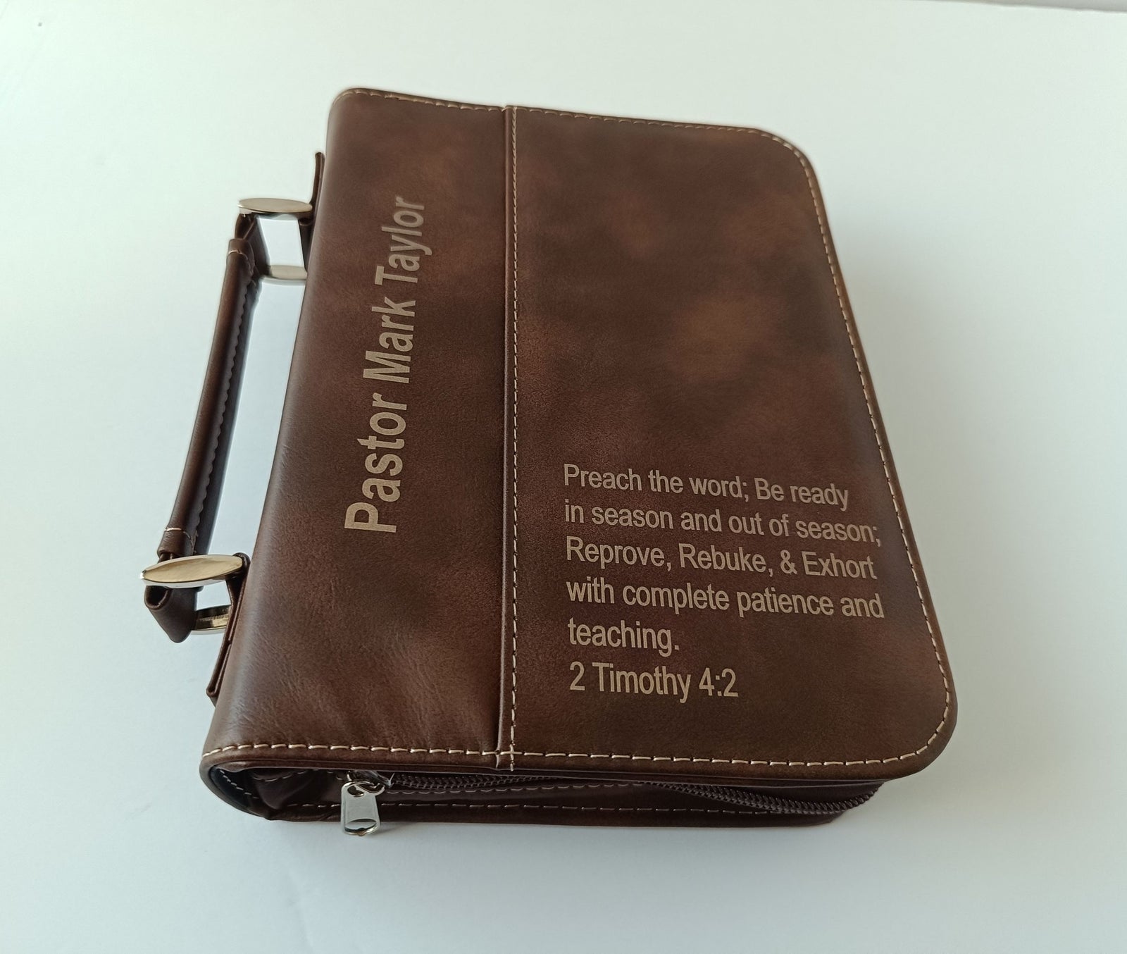 A brown leather Bible or book cover with a handle, featuring the name 'Pastor Mark Taylor' on the left and a Bible verse from 2 Timothy 4:2 on the right, photographed on a white background.