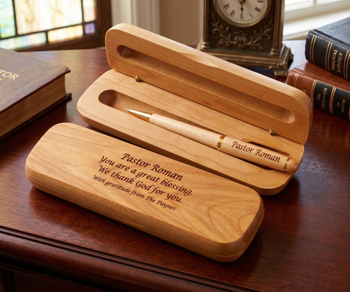 A personalized wooden pen gift set displayed on a dark wood desk, featuring an open wooden case with a white and gold pen inside, alongside a closed matching wooden case engraved with the name 'Pastor Roman' and a thank you message.