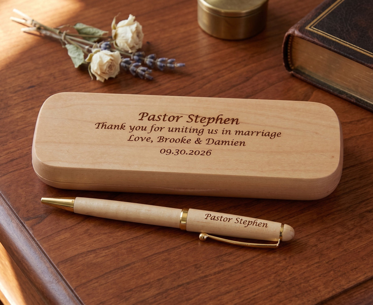 Personalized wooden wedding keepsake box and matching pen engraved with 'Pastor Stephen' and wedding details, displayed on a wooden table with dried flowers and a book in the background.