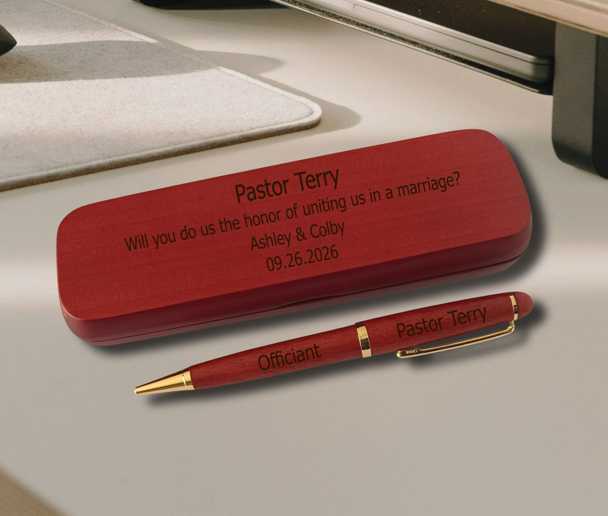 A personalized red wooden wedding keepsake box and matching pen set engraved with 'Pastor Terry' and wedding details, displayed on a neutral desk surface.