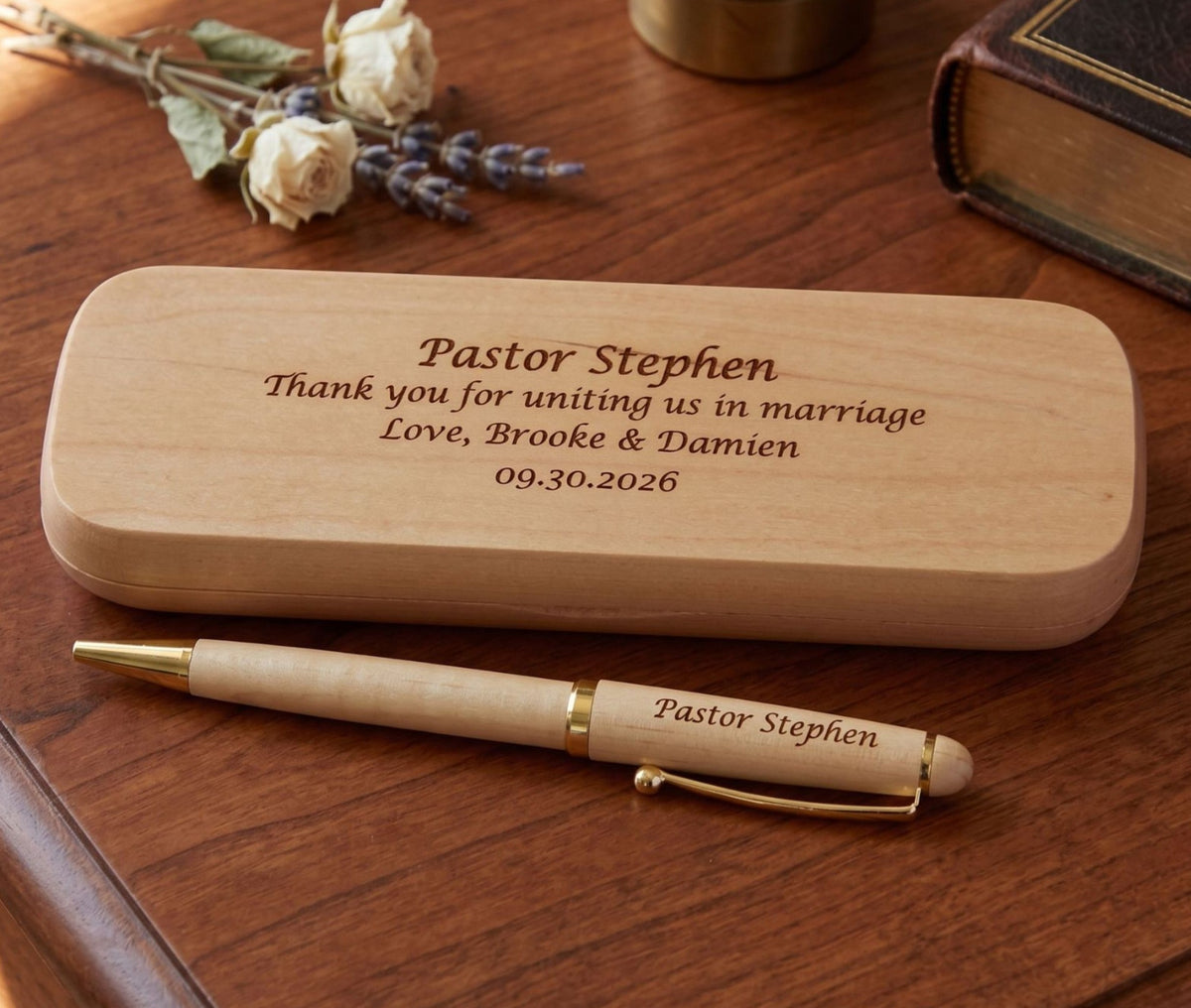 Personalized wooden wedding keepsake box and matching pen engraved with 'Pastor Stephen' and wedding details, displayed on a wooden table with dried flowers and a book in the background.