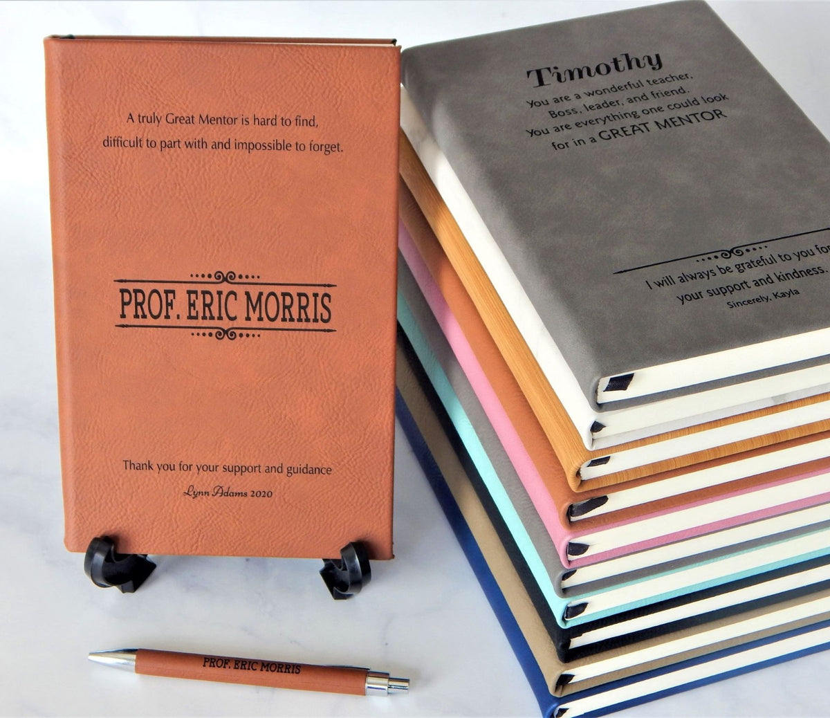 Personalized leather journal covers in multiple colors, including a prominent terracotta orange one standing upright and a stack of notebooks in gray, peach, orange, pink, blue, and green. A matching terracotta orange pen lies at the bottom on a white surface.