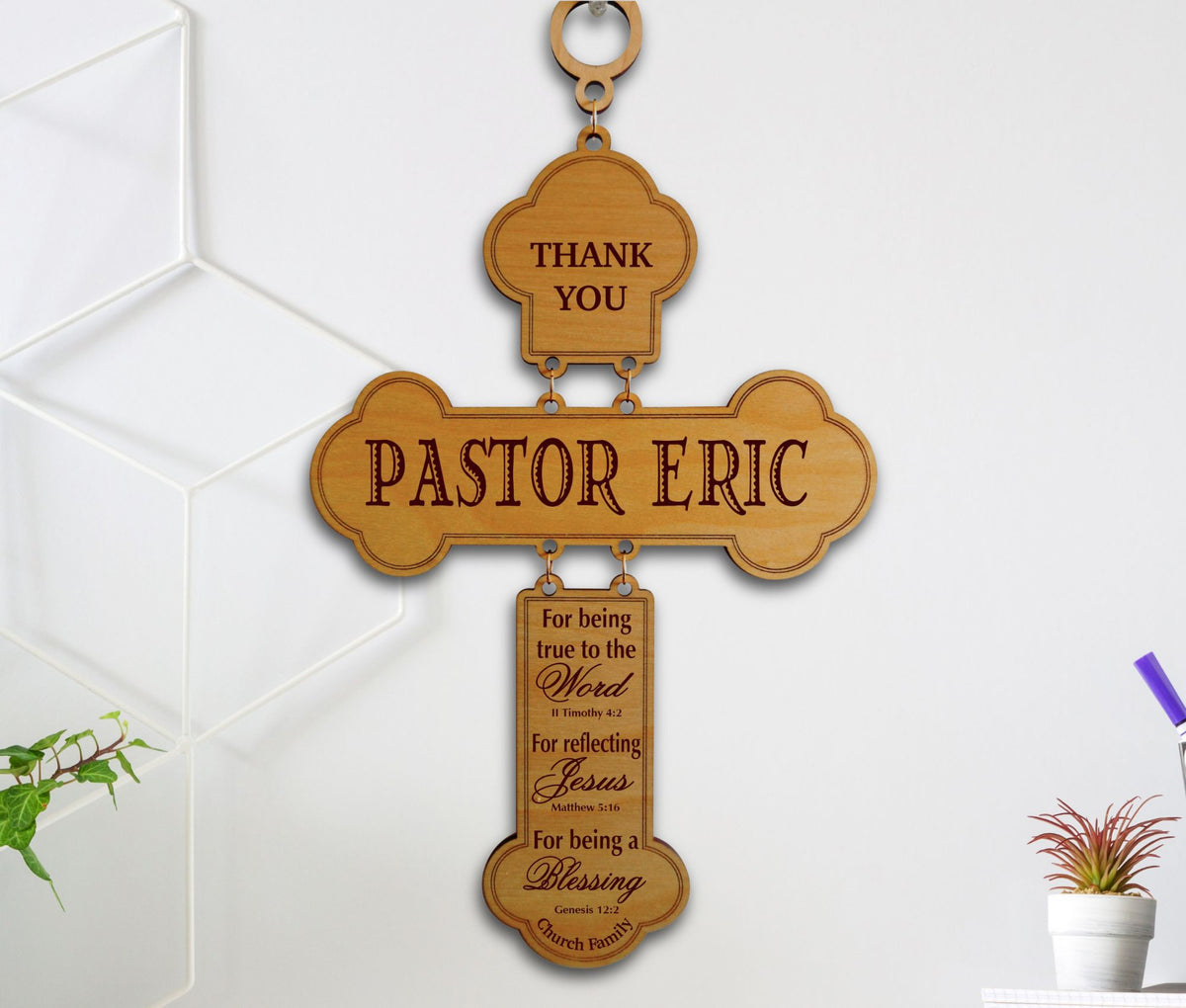 A personalized wooden cross wall hanging with engraved text reading 'THANK YOU PASTOR ERIC' and a hanging plaque with scripture, shown against a white wall with decorative wall panels and small plants.