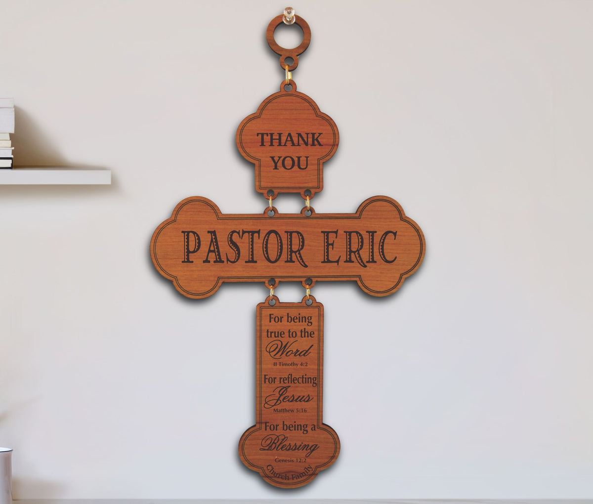 A wooden cross-shaped wall sign with engraved text reading 'THANK YOU PASTOR ERIC' and additional scripture-style text on a hanging tab, mounted on a light wall.