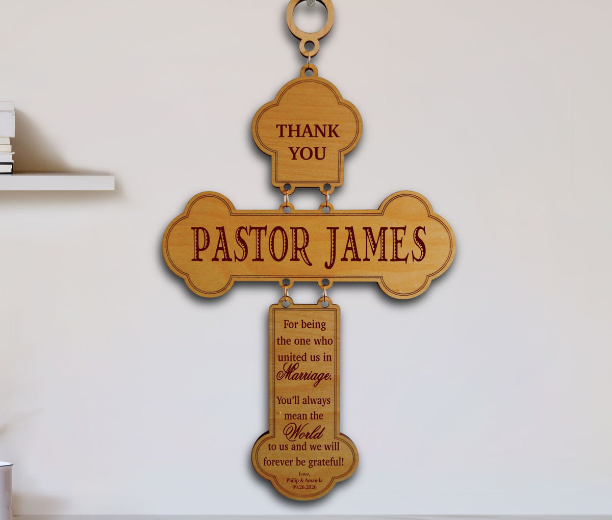 A personalized wooden cross wall sign with engraved text reading 'THANK YOU PASTOR JAMES' and a blessing below, hanging against a white wall.