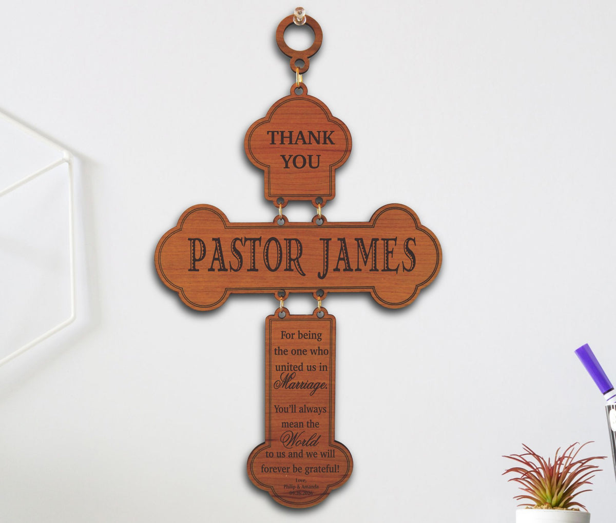 A personalized wooden cross wall hanging with engraved text reading 'THANK YOU' and 'PASTOR JAMES' on a light gray wall.
