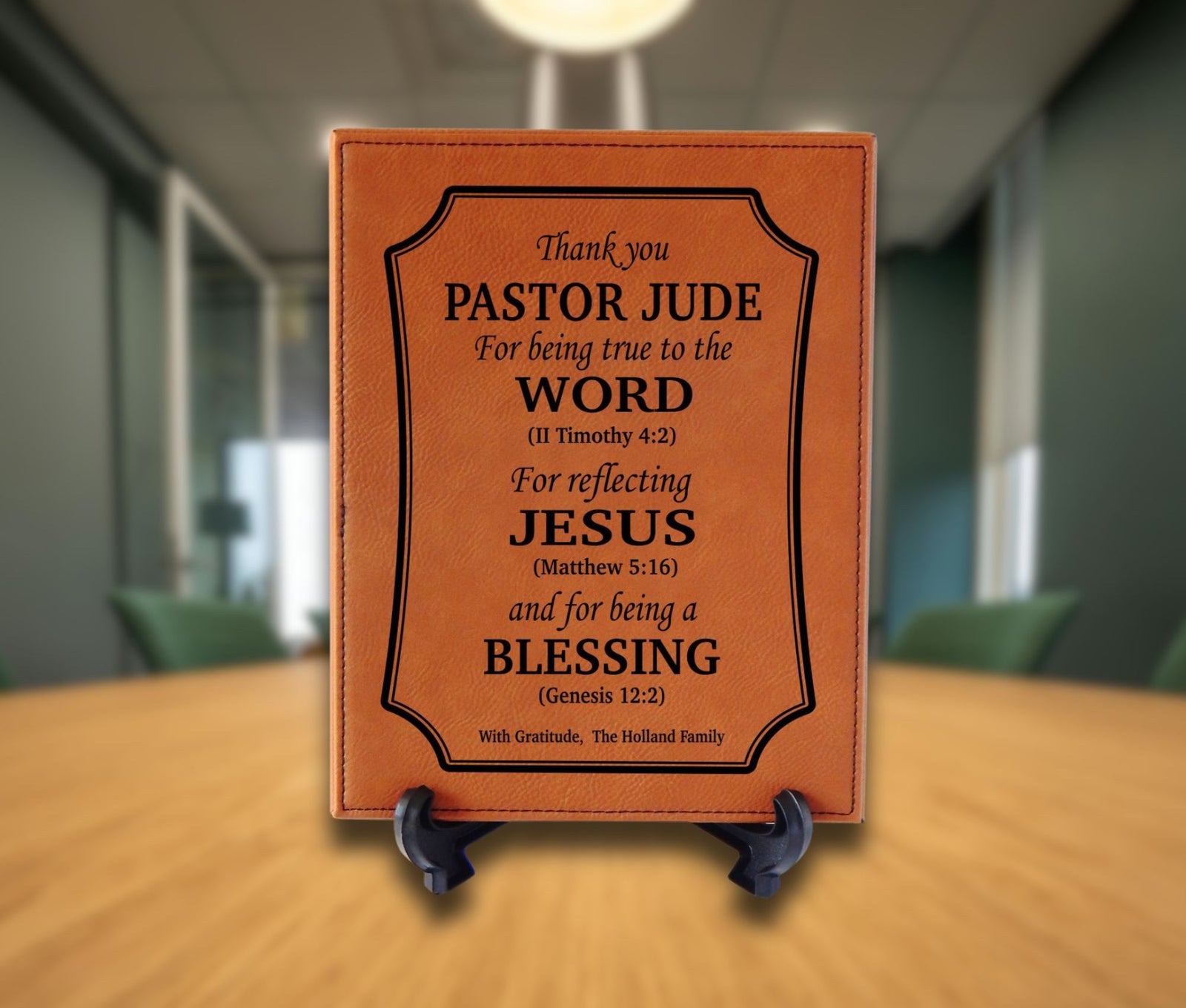 A rectangular brown leather plaque with black printed text, displayed on a small black stand on a wooden table in a blurred office setting.