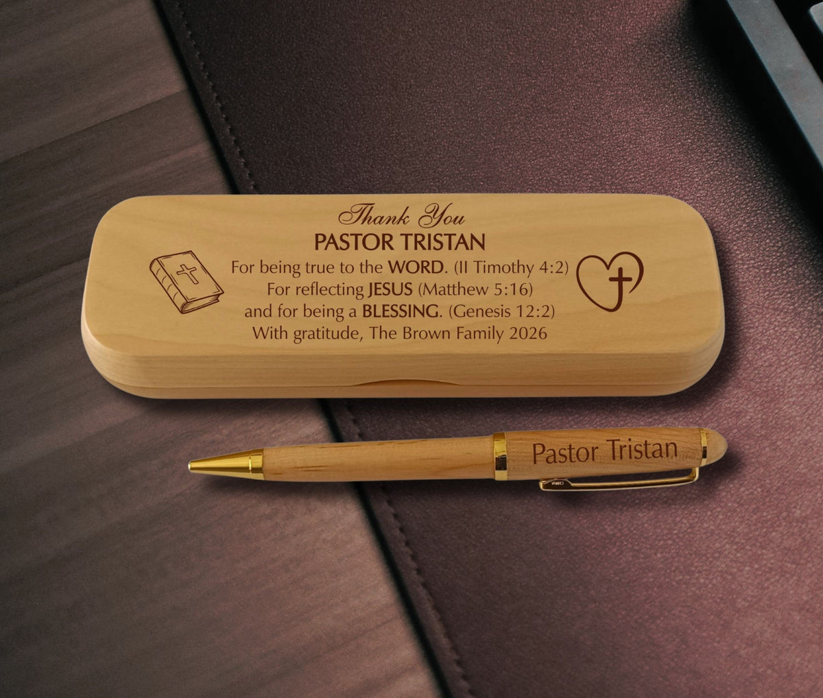 A personalized wooden pen with gold trim and a matching wooden keepsake box, both engraved with the name 'PASTOR TRISTAN' and a Bible verse, displayed on a dark brown leather surface.