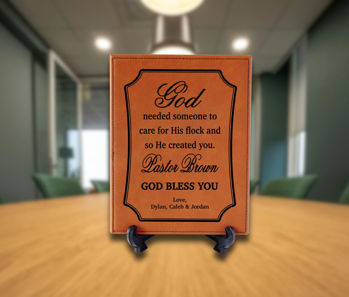 A square brown leather-bound plaque with black engraved text stands on a small black easel on a wooden table. The background shows a softly blurred office or conference room interior with chairs and ceiling lights.