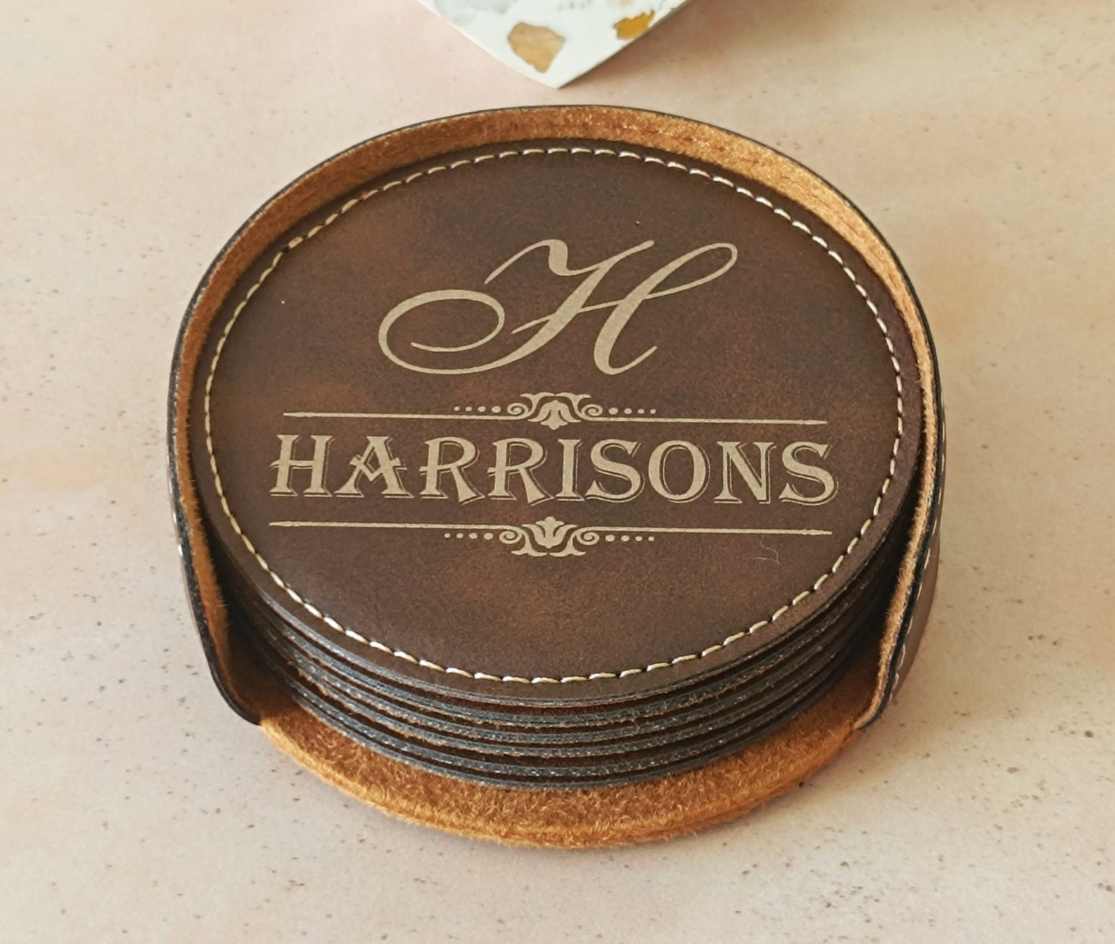 A stack of round brown leather coasters with white stitching, featuring a top coaster engraved with a large script letter H and the name HARRISONS in serif text, shown on a light beige surface.