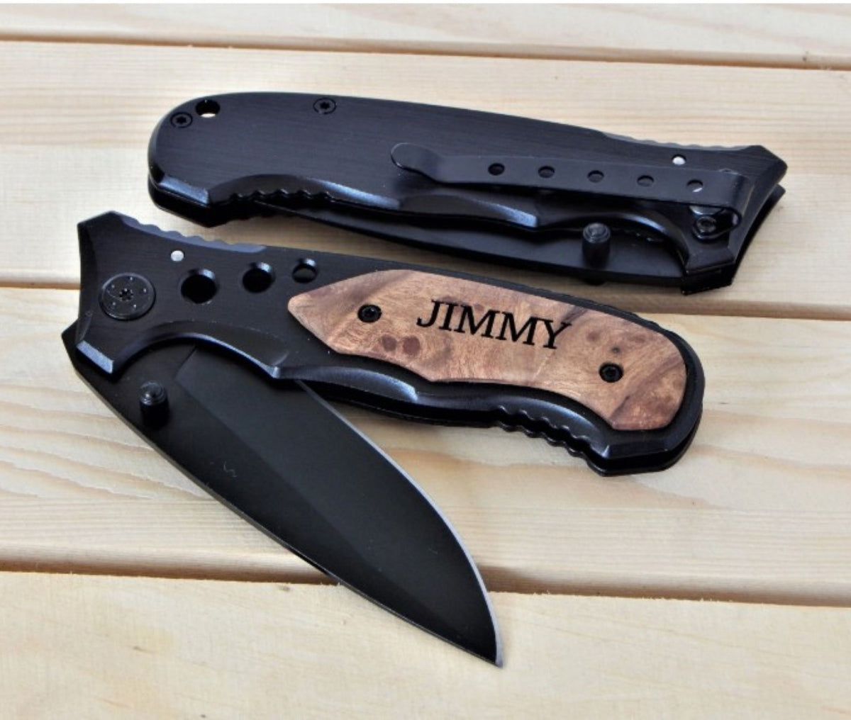 Two folding pocket knives displayed on a light wood plank surface. One knife is open, showing a matte black blade and a black handle with a natural wood inlay engraved with the name 'JIMMY'.