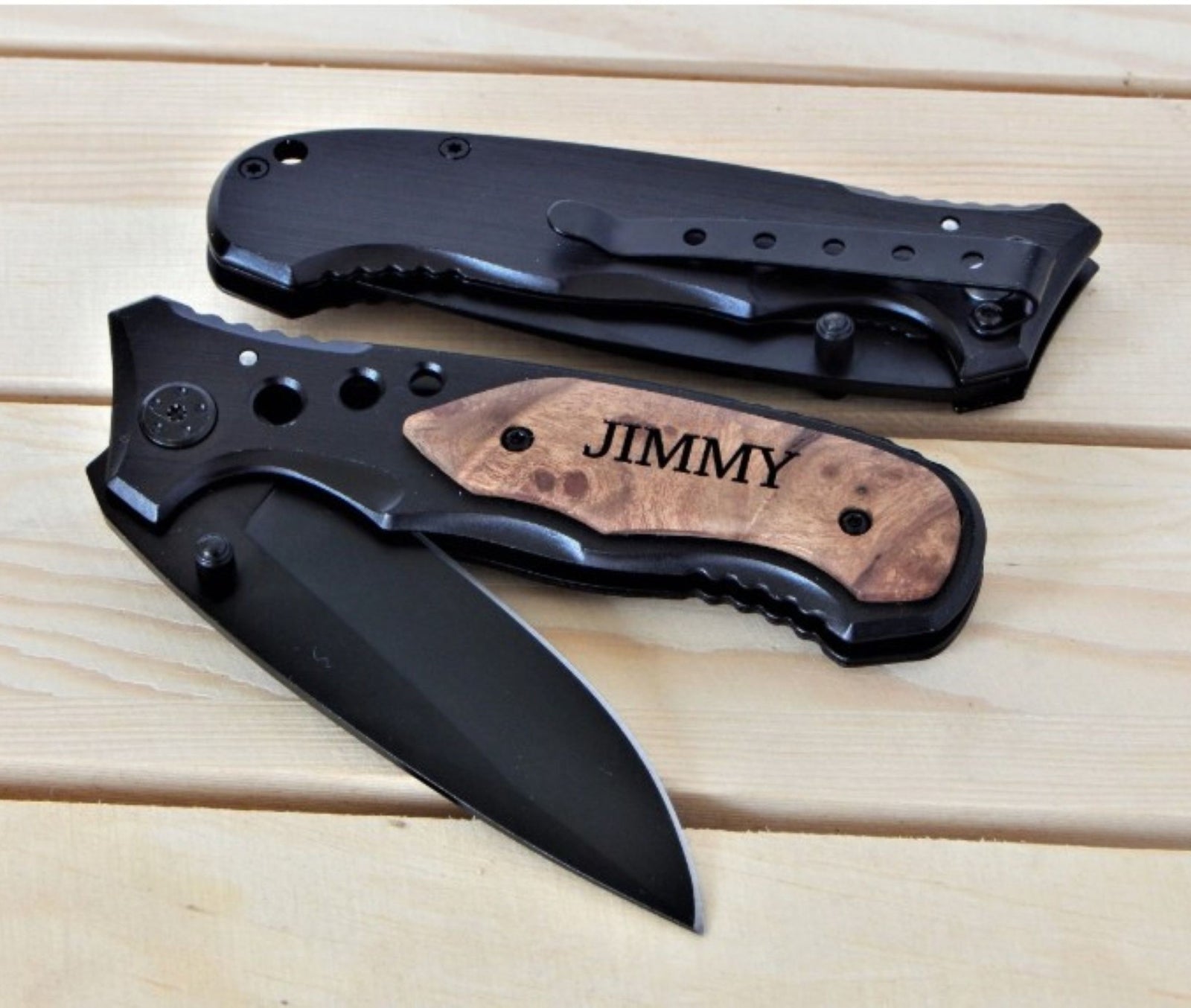 Two folding pocket knives displayed on a light wood plank surface. One knife is open, showing a matte black blade and a black handle with a natural wood inlay engraved with the name 'JIMMY'.